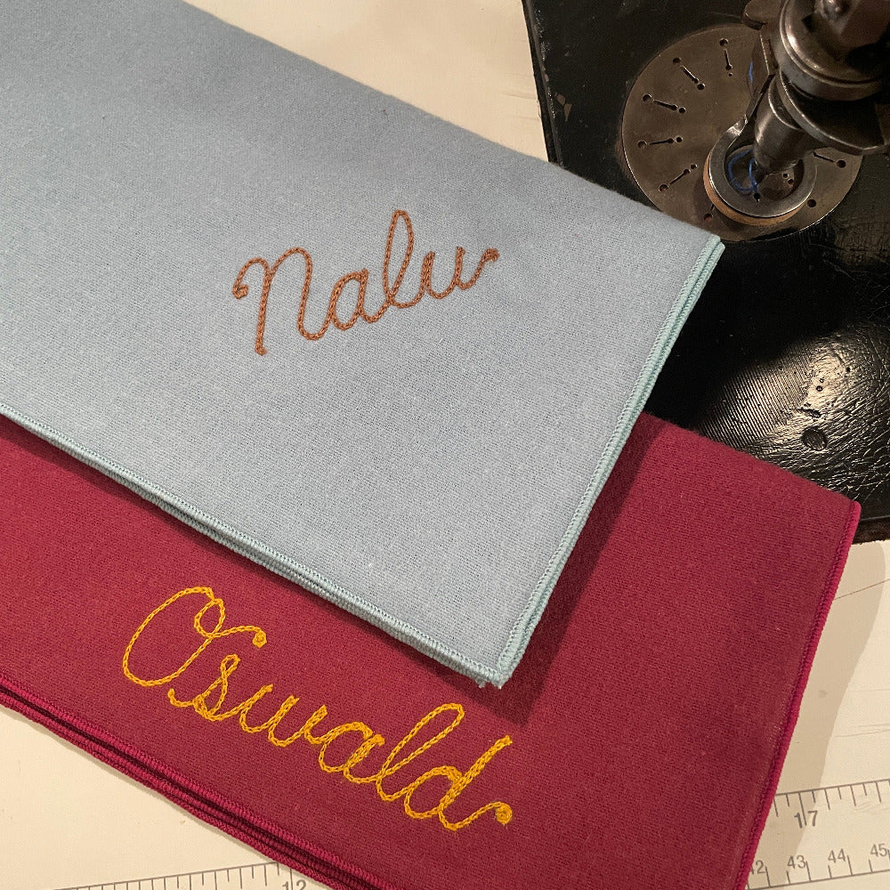 Two Dog Bandanas with chain stitched embroidered names on a surface with a chain stitching machine in the background